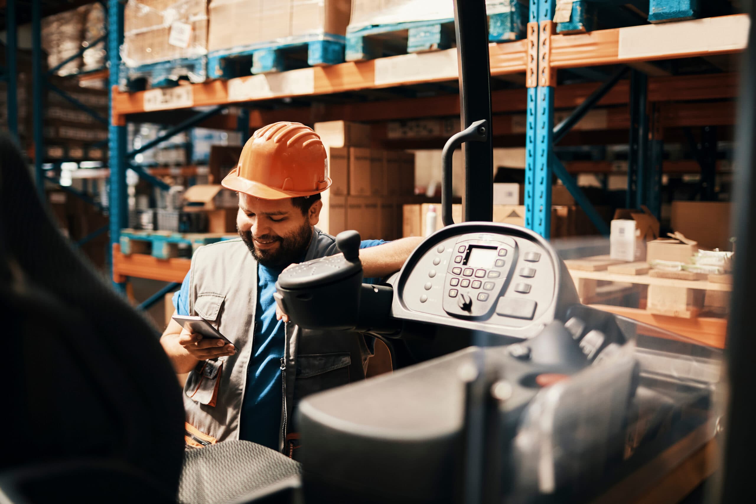 Smiling male warehouse worker in hard hat using smartphone next to forklift