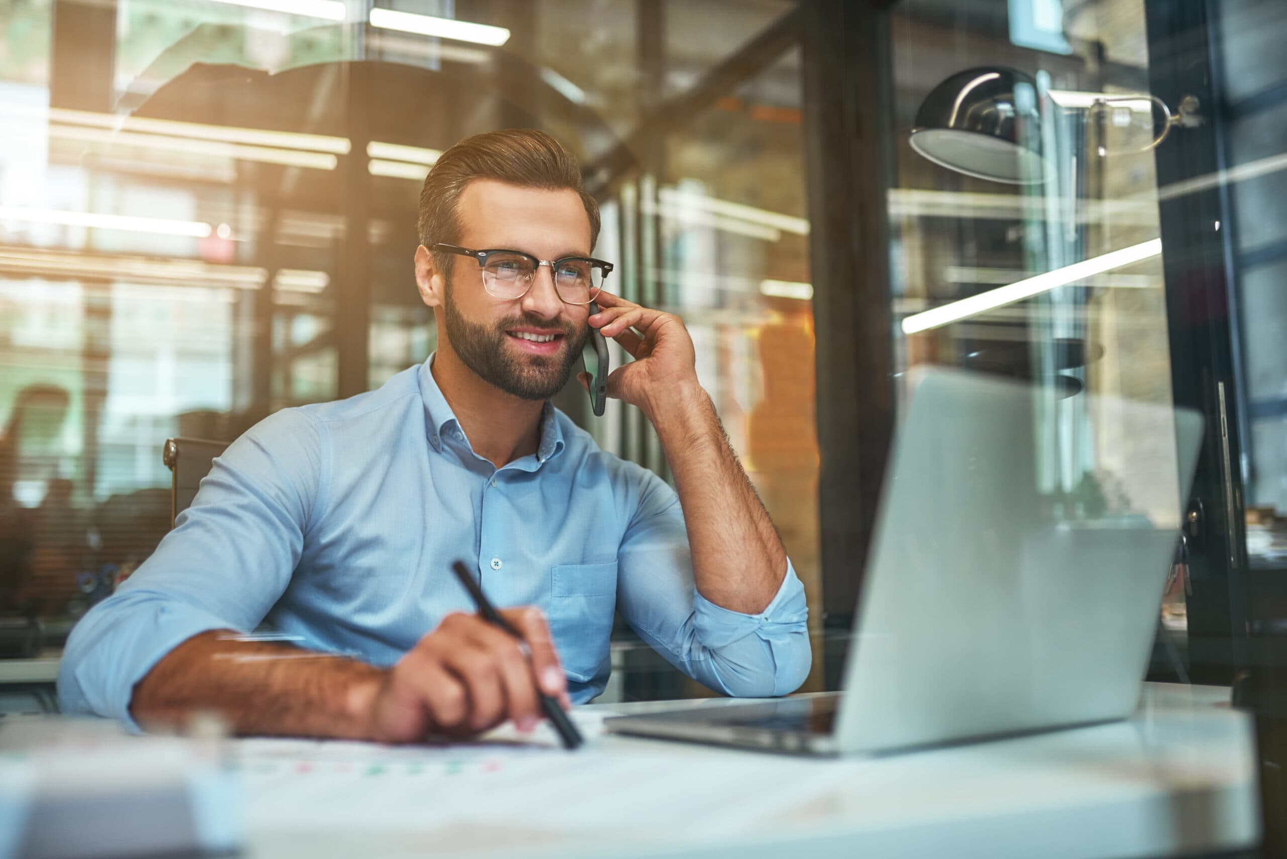 Customer support. Portrait of young and cheerful businessman in eyeglasses and formal wear talking on the phone and smiling while sitting in the office. Working place. Business concept